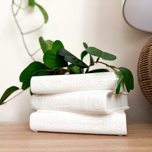 May include: A white ceramic book-shaped planter with the titles "Wuthering Heights", "Emma", and "Little Women" embossed on the spines. The planter holds green foliage, creating a decorative display.
