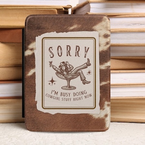 Op de afbeelding: Een bruin en wit patroonhoesje met de tekst "SORRY I'M BUSY DOING COWGIRL STUFF RIGHT NOW". De hoes heeft een illustratie van een cowgirl die ontspant in een cocktailglas. De hoes is omgeven door een stapel boeken.