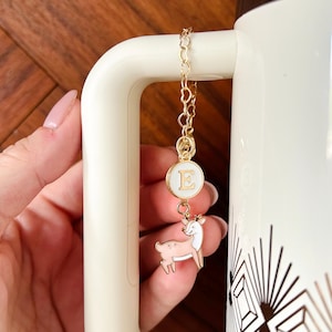 May include: A gold-colored chain charm with a white circular pendant featuring the letter "E" and a small pink and white deer charm. The charm is attached to a white mug.