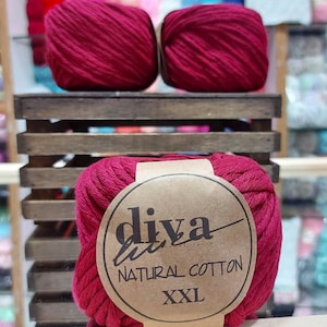 May include: Three skeins of deep red Diva Natural Cotton XXL yarn are displayed on a wooden shelf. The label on the bottom skein is visible, showcasing the brand and product details. The yarn is tightly wound, suggesting a high-quality crafting material.
