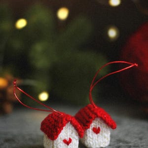 May include: Two small, handmade crochet ornaments shaped like houses. Each house has a white body with a small red heart embroidered on the front and a textured red roof. A thin red string is attached to the top of each for hanging.