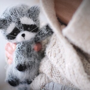 May include: A small, fuzzy, handcrafted raccoon figurine made of grey, white, and black yarn. The raccoon features a distinct black mask around its eyes, pointed ears, a small black nose, and long white whiskers extending from its snout.