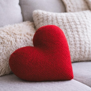 Könnte beinhalten: Ein rotes, gestricktes herzförmiges Kissen liegt auf einem grauen Sofa, umgeben von anderen Kissen und einer Decke. Das Herzkissen ist der Blickfang, mit einer strukturierten Oberfläche. Weiche Beleuchtung und gemütliche Atmosphäre.