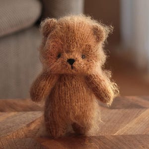 May include: A small, fuzzy, light brown knitted bear figurine with a soft, textured appearance. The bear features rounded ears, a dark brown stitched nose, and small dark eyes, standing upright with a simple, compact body shape.