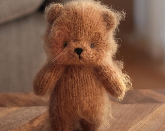 May include: A small, fuzzy, light brown knitted bear figurine with a soft, textured appearance. The bear features rounded ears, a dark brown stitched nose, and small dark eyes, standing upright with a simple, compact body shape.