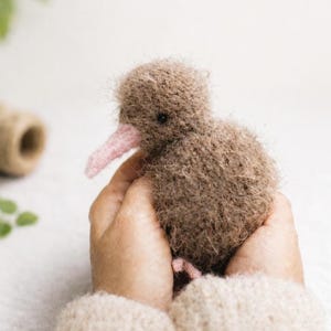 May include: A handcrafted brown kiwi bird soft toy with a pink beak is held in two hands. The bird has a textured, fuzzy appearance. The hands are wearing a beige jumper. The background is a soft, neutral colour.