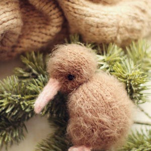 K&ouml;nnte beinhalten: Ein handgefertigter, brauner Kiwi-Vogel aus Pl&uuml;sch mit rosa Schnabel und F&uuml;&szlig;en, der auf gr&uuml;nen Kiefernzweigen ruht. Der Vogel ist aus weichem, flauschigem Material gefertigt. Eine beige Strickdecke befindet sich im Hintergrund.