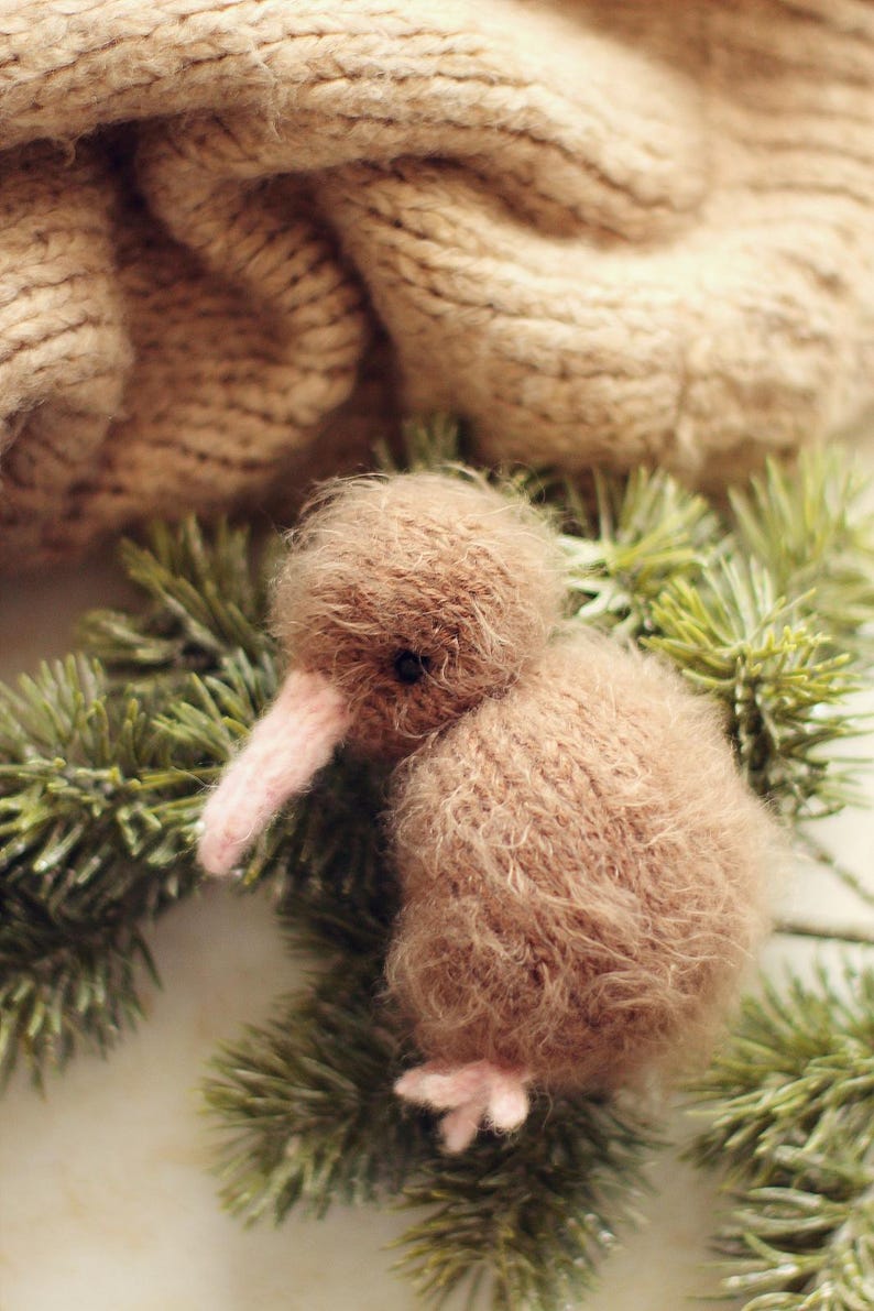 K&ouml;nnte beinhalten: Ein handgefertigter, brauner Kiwi-Vogel aus Pl&uuml;sch mit rosa Schnabel und F&uuml;&szlig;en, der auf gr&uuml;nen Kiefernzweigen ruht. Der Vogel ist aus weichem, flauschigem Material gefertigt. Eine beige Strickdecke befindet sich im Hintergrund.