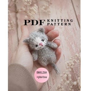 Op de afbeelding: Een klein, handgebreid grijs en wit kitten-speeltje met een roze neus en zwarte snorharen. De afbeelding bevat de tekst PDF KNITTING PATTERN en een roze cirkel met de tekst ENGLISH +photos, wat duidt op een digitaal instructiedocument.