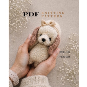 May include: A PDF knitting pattern document featuring a small, hand-knitted cream-colored plush puppy with long, textured brown ears and a small beige bow on its head. The text on the document reads PDF KNITTING PATTERN and ENGLISH +photos.