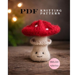 Könnte beinhalten: Ein gestrickter Pilz mit rotem Hut und weißen Punkten, einem lächelnden Gesicht und einem beigen Stiel. Das Bild enthält den Text "PDF KNITTING PATTERN" und "ENGLISH + photos". Der Pilz steht auf einer Holzoberfläche.
