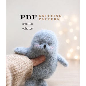 May include: A handmade, light blue knitted seal toy with rosy cheeks and small black eyes. The toy is held by a person wearing a beige sweater. Text on the image reads "PDF KNITTING PATTERN ENGLISH +photos".