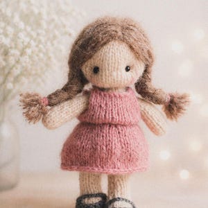 May include: A handmade knitted doll with brown yarn hair styled in two plaits. The doll wears a pink dress and grey shoes. The doll's arms and legs are beige. The background is a soft, blurred neutral colour.