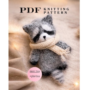 May include: A PDF knitting pattern for a small, fuzzy gray and white raccoon toy. The raccoon has black eye patches, a black nose, and wears a cream-colored knitted scarf. The text reads PDF KNITTING PATTERN and ENGLISH +photos.