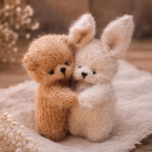Peut inclure: Deux peluches tricotées, une beige et une crème, s'enlacent. Le jouet beige a la forme d'un ours en peluche, tandis que le jouet crème a de longues oreilles de lapin. Elles sont posées sur un tissu de ton neutre, créant une scène chaleureuse.