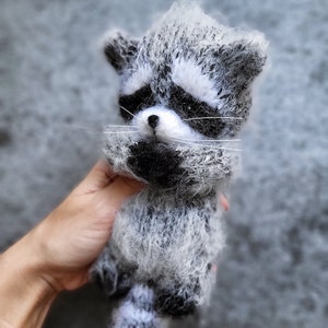 May include: A fuzzy, handcrafted raccoon figurine made of textured grey, white, and black yarn. The raccoon features a distinct black mask around its eyes, a small black nose, long white whiskers, and a striped tail with alternating grey and white bands.