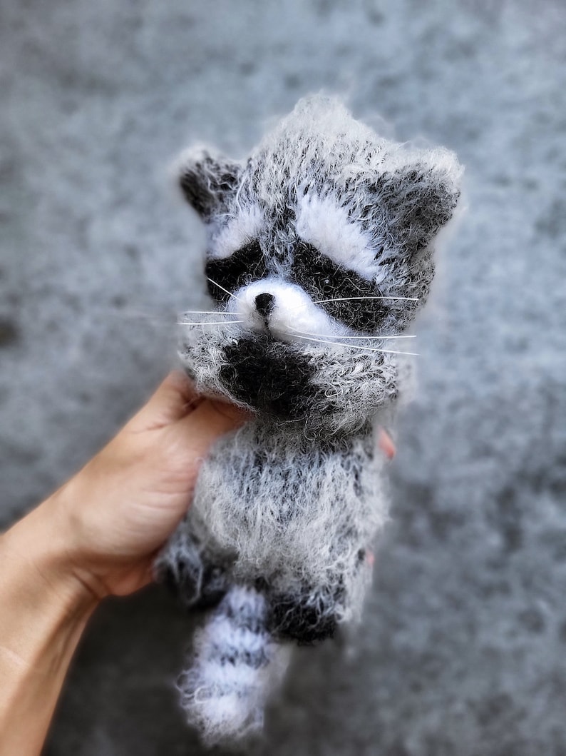 May include: A fuzzy, handcrafted raccoon figurine made of textured grey, white, and black yarn. The raccoon features a distinct black mask around its eyes, a small black nose, long white whiskers, and a striped tail with alternating grey and white bands.