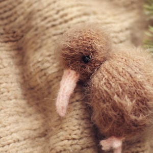 K&ouml;nnte beinhalten: Ein handgefertigtes braunes Kiwi-Vogel-Pl&uuml;schtier mit einem langen rosa Schnabel und kleinen rosa F&uuml;&szlig;en. Der Vogel besteht aus weichem, flauschigem Material und liegt auf einer beigefarbenen Strickoberfl&auml;che. Eine Garnrolle ist im Hintergrund zu sehen.