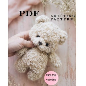 May include: A PDF knitting pattern for a small, beige, fuzzy teddy bear. The document includes text that reads PDF KNITTING PATTERN and a pink circular badge stating ENGLISH +photos. The bear has dark eyes and a dark nose.