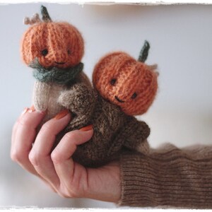 Op de afbeelding: Twee gebreide pompoenfiguurtjes met lachende gezichten. De pompoenen zijn oranje en hebben groene stelen. De figuurtjes worden vastgehouden in een hand die een bruine trui draagt.