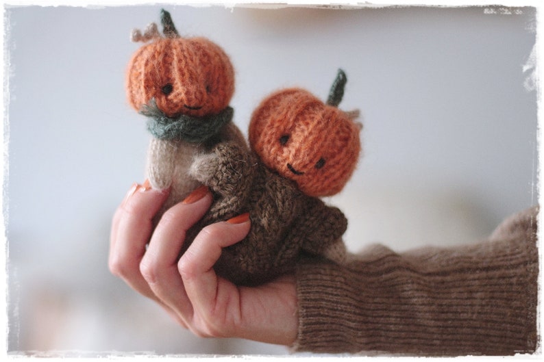 Op de afbeelding: Twee gebreide pompoenfiguurtjes met lachende gezichten. De pompoenen zijn oranje en hebben groene stelen. De figuurtjes worden vastgehouden in een hand die een bruine trui draagt.