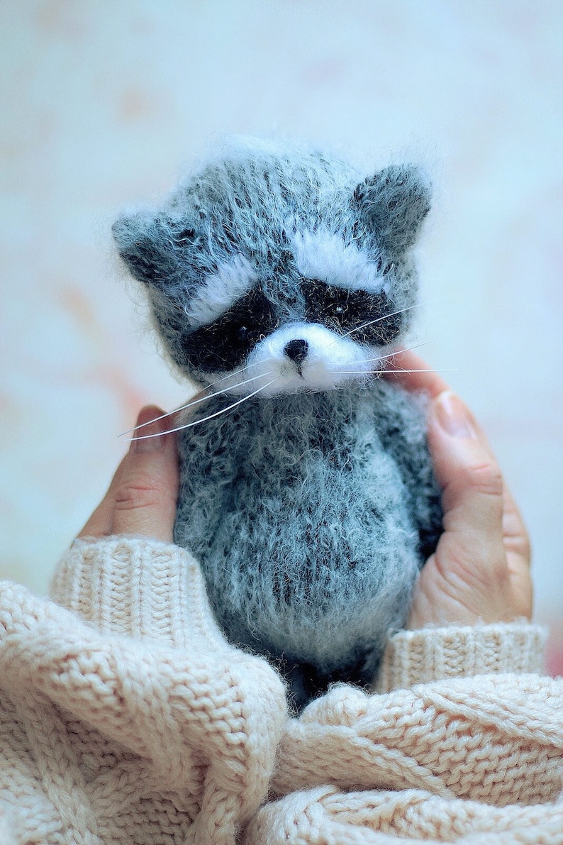 May include: A small, fuzzy, handcrafted raccoon figurine made of grey and white textured yarn. The raccoon features a distinct black mask around its dark eyes, a small black nose, and long, thin white whiskers extending from its snout.