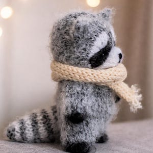 May include: A small, fuzzy, hand-knitted raccoon figurine made of grey and white yarn. The raccoon features a black eye mask, a striped tail, and is wearing a cream-coloured knitted scarf wrapped around its neck. The texture is soft and fibrous.