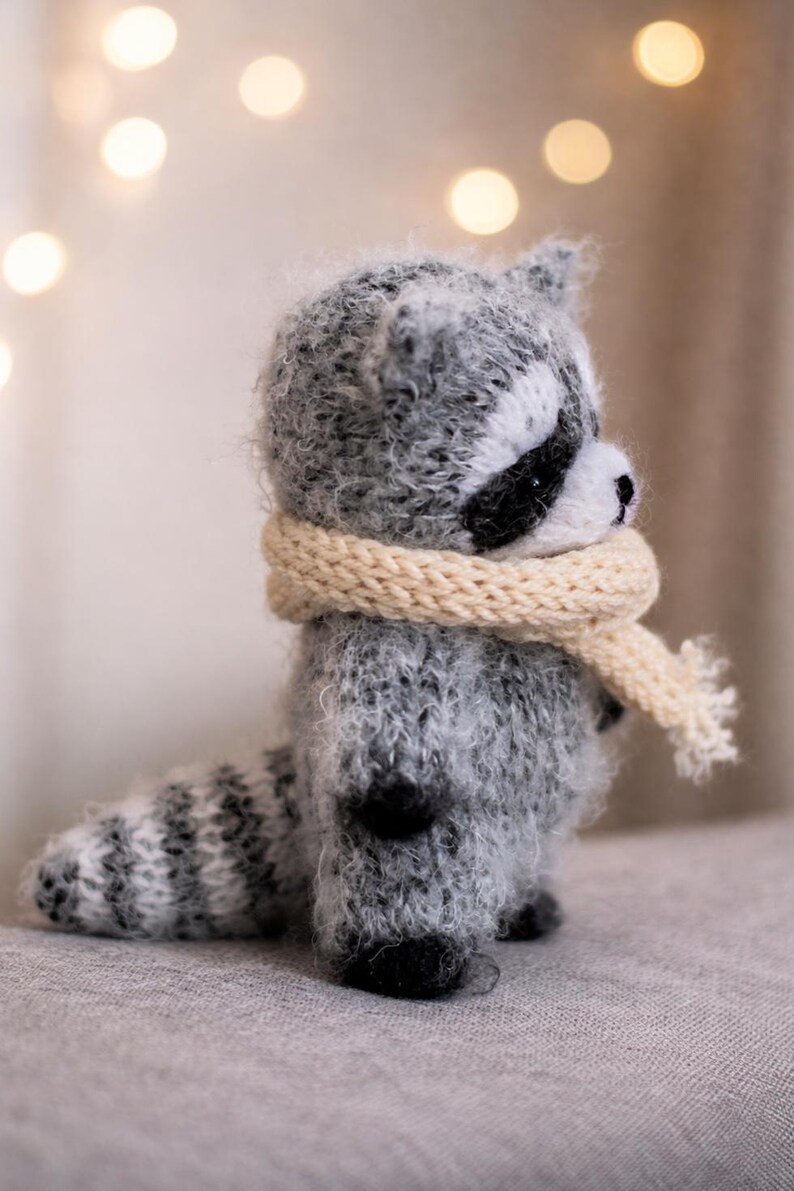 May include: A small, fuzzy, hand-knitted raccoon figurine made of grey and white yarn. The raccoon features a black eye mask, a striped tail, and is wearing a cream-coloured knitted scarf wrapped around its neck. The texture is soft and fibrous.
