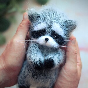 May include: A small, fuzzy, handcrafted raccoon figurine made of grey, white, and black yarn. The raccoon features a distinct black eye mask, a white muzzle with a small black nose, and long, thin white whiskers extending from its cheeks.