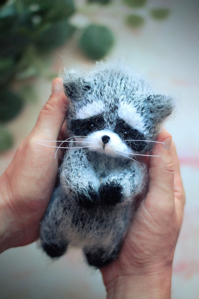 May include: A small, fuzzy, handcrafted raccoon figurine made of grey, white, and black yarn. The raccoon features a distinct black eye mask, a white muzzle with a small black nose, and long, thin white whiskers extending from its cheeks.