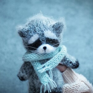 May include: A small, fuzzy, handcrafted raccoon figurine made of grey and white yarn. The raccoon features a black mask around its eyes, a small black nose, and long white whiskers. It is wearing a light blue knitted scarf with fringed ends.