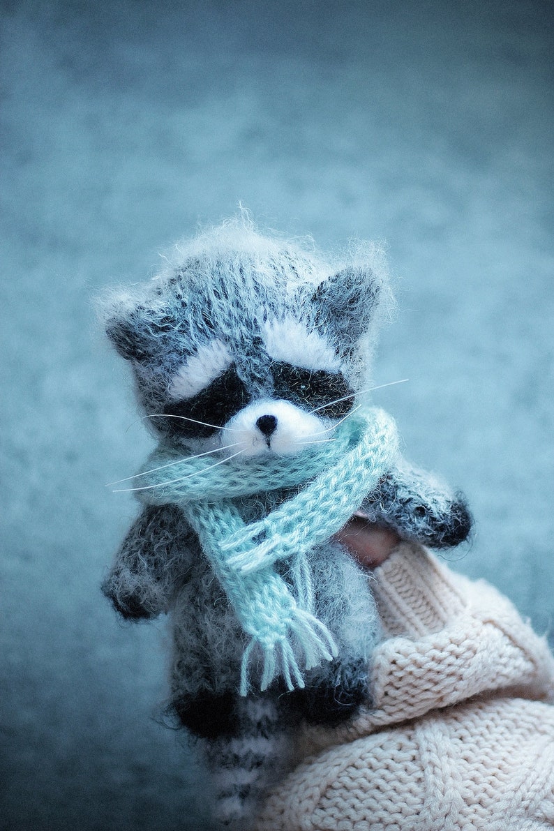 May include: A small, fuzzy, handcrafted raccoon figurine made of grey and white yarn. The raccoon features a black mask around its eyes, a small black nose, and long white whiskers. It is wearing a light blue knitted scarf with fringed ends.