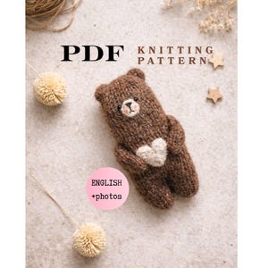 May include: A PDF knitting pattern for a small brown knitted bear holding a cream-colored heart. The text reads PDF KNITTING PATTERN and ENGLISH +photos. The bear has embroidered facial features and is surrounded by two small pom-poms and two stars.