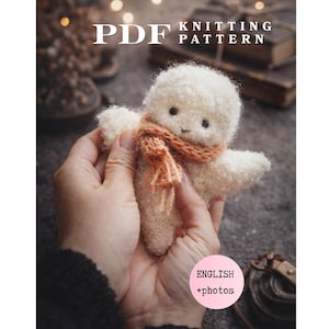 Könnte beinhalten: Eine PDF-Strickanleitung mit einer kleinen, flauschigen, cremefarbenen gehäkelten Figur mit schwarzen Perlenaugen und einem kleinen orangefarbenen Schal. Der Text auf dem Bild lautet PDF KNITTING PATTERN und ENGLISH +photos in einem rosa Kreis.