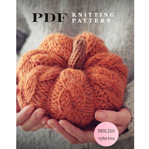May include: A PDF knitting pattern document featuring a hand-knit orange pumpkin with a brown stem. The text on the document reads PDF KNITTING PATTERN and ENGLISH +photos. The pumpkin has a textured cable knit design and a rounded shape.