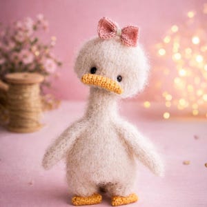 Może przedstawiać: Ręcznie robiona, puszysta, biała pluszowa kaczka z różową kokardą na głowie. Kaczka ma pomarańczowy dziób i stopy oraz małe czarne oczy. Tło jest w kolorze miękkiego różu ze światłami bokeh, sugerującym kapryśny motyw.