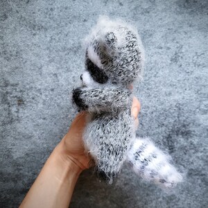 May include: A small, fuzzy, hand-knitted raccoon figurine crafted from grey, white, and black yarn. The plush toy features a distinct striped tail, pointed ears, and a dark mask pattern around the eyes, showcasing a soft, textured, and fibrous appearance.