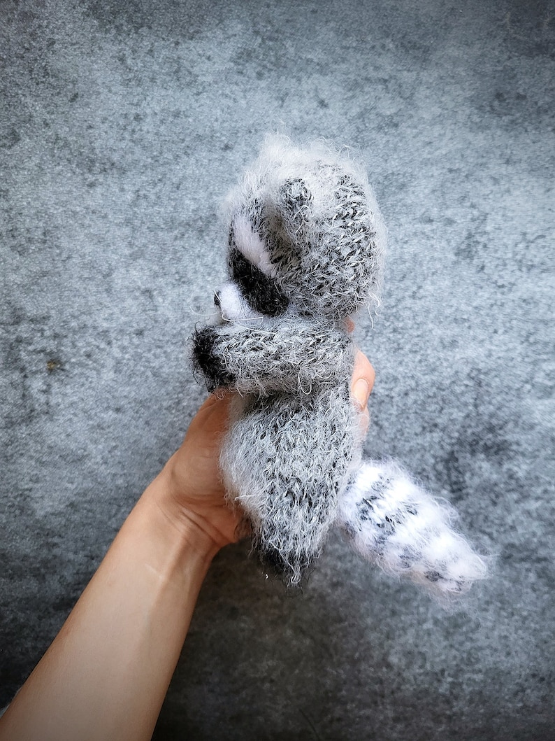 May include: A small, fuzzy, hand-knitted raccoon figurine crafted from grey, white, and black yarn. The plush toy features a distinct striped tail, pointed ears, and a dark mask pattern around the eyes, showcasing a soft, textured, and fibrous appearance.
