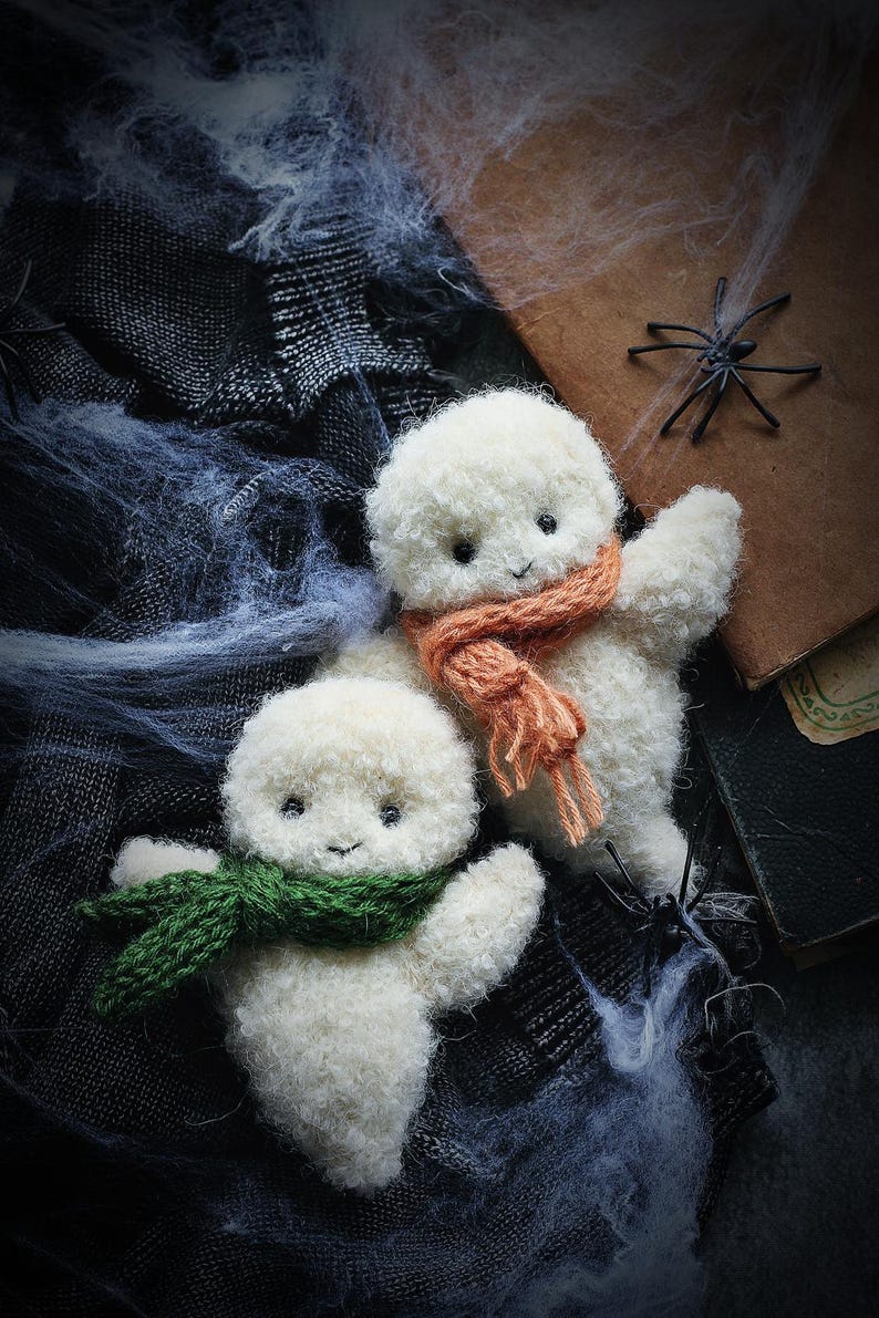 K&ouml;nnte beinhalten: Zwei flauschige, wei&szlig;e Geisterfiguren mit orangefarbenen und gr&uuml;nen Schals, platziert vor einem Hintergrund aus Spinnweben und dunklem Stoff. Eine kleine schwarze Spinne und ein Vintage-Buch erzeugen eine Halloween-Atmosph&auml;re.