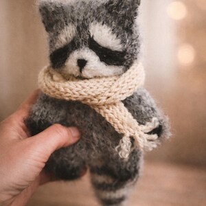 May include: A small, fuzzy, hand-knitted raccoon toy made of grey and white yarn. The raccoon features a distinct black mask around its eyes, a small black nose, and a striped tail. It is wearing a thick, cream-coloured knitted scarf around its neck.