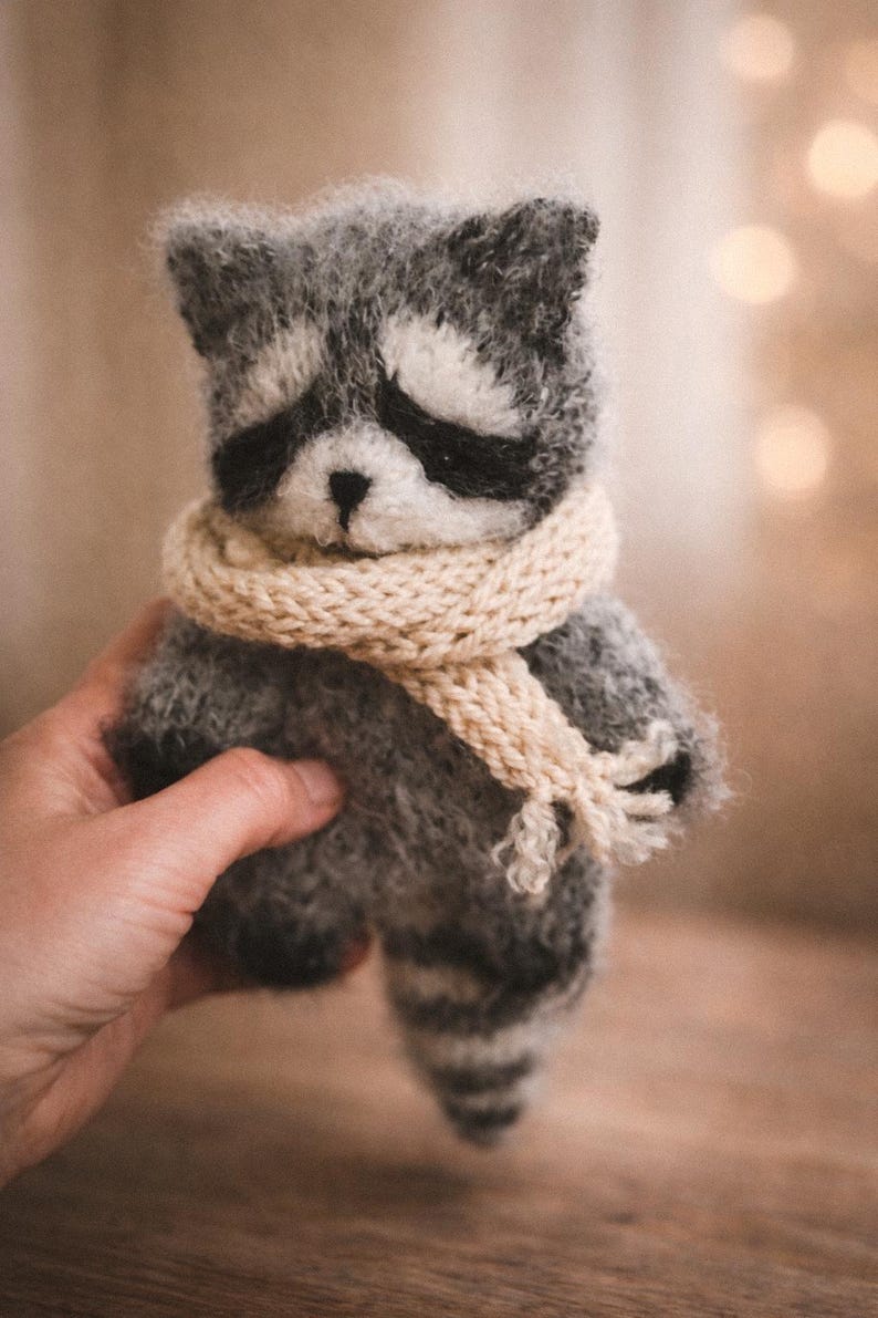 May include: A small, fuzzy, hand-knitted raccoon toy made of grey and white yarn. The raccoon features a distinct black mask around its eyes, a small black nose, and a striped tail. It is wearing a thick, cream-coloured knitted scarf around its neck.