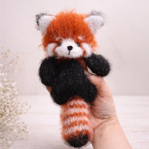 May include: A handmade red panda plush toy with orange, white, black, and grey details. The plush has a fluffy orange head, black arms and a striped tail. The toy is held in a hand.