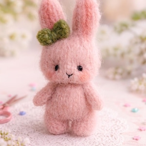 Puede incluir: Una figura de conejo rosa hecha a mano con un lazo verde. El conejo tiene ojos negros y mejillas rosadas. Está sobre un mantel individual de encaje blanco. El fondo es suave y borroso.