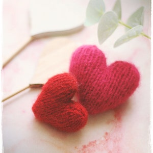 Puede incluir: Dos corazones tejidos, uno rojo y otro rosa, se colocan sobre una superficie blanca con un fondo rosa claro. Los corazones están hechos de hilo y tienen una textura suave y peluda.
