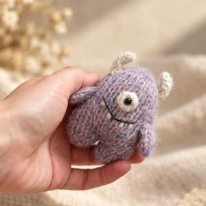 Puede incluir: Un pequeño juguete de monstruo tejido a mano con hilo jaspeado en tonos morados y azules. La criatura presenta dos pequeños cuernos de color crema, un solo ojo central con pupila negra y una boca negra bordada con un diminuto diente blanco.
