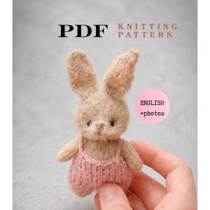 May include: A small, tan-colored knitted bunny wearing a pink overall. The image includes the text "PDF KNITTING PATTERN" and "ENGLISH +photos". The bunny is held in a hand against a gray background.
