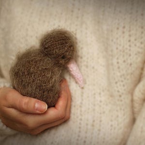 K&ouml;nnte beinhalten: Ein handgefertigtes braunes Kiwi-Vogel-Pl&uuml;schtier mit einem rosa Schnabel wird in einer Hand gehalten. Der Vogel besteht aus einem weichen, strukturierten Material. Der Hintergrund ist ein cremefarbener Pullover.