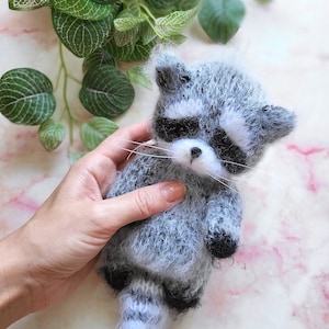 May include: A small, fuzzy, handcrafted raccoon figurine made of grey and white yarn. The raccoon features a distinct black mask around its eyes, a white muzzle with a small black nose, long white whiskers, and a striped tail with black paws.