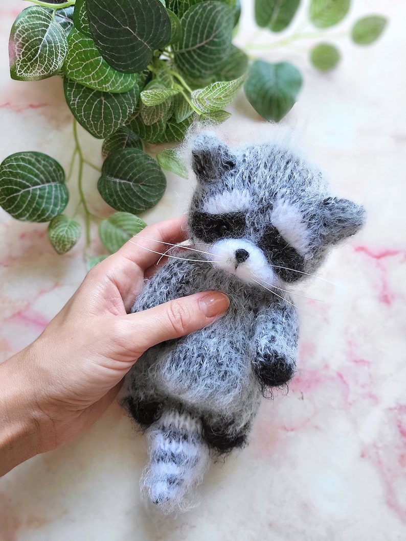 May include: A small, fuzzy, handcrafted raccoon figurine made of grey and white yarn. The raccoon features a distinct black mask around its eyes, a white muzzle with a small black nose, long white whiskers, and a striped tail with black paws.