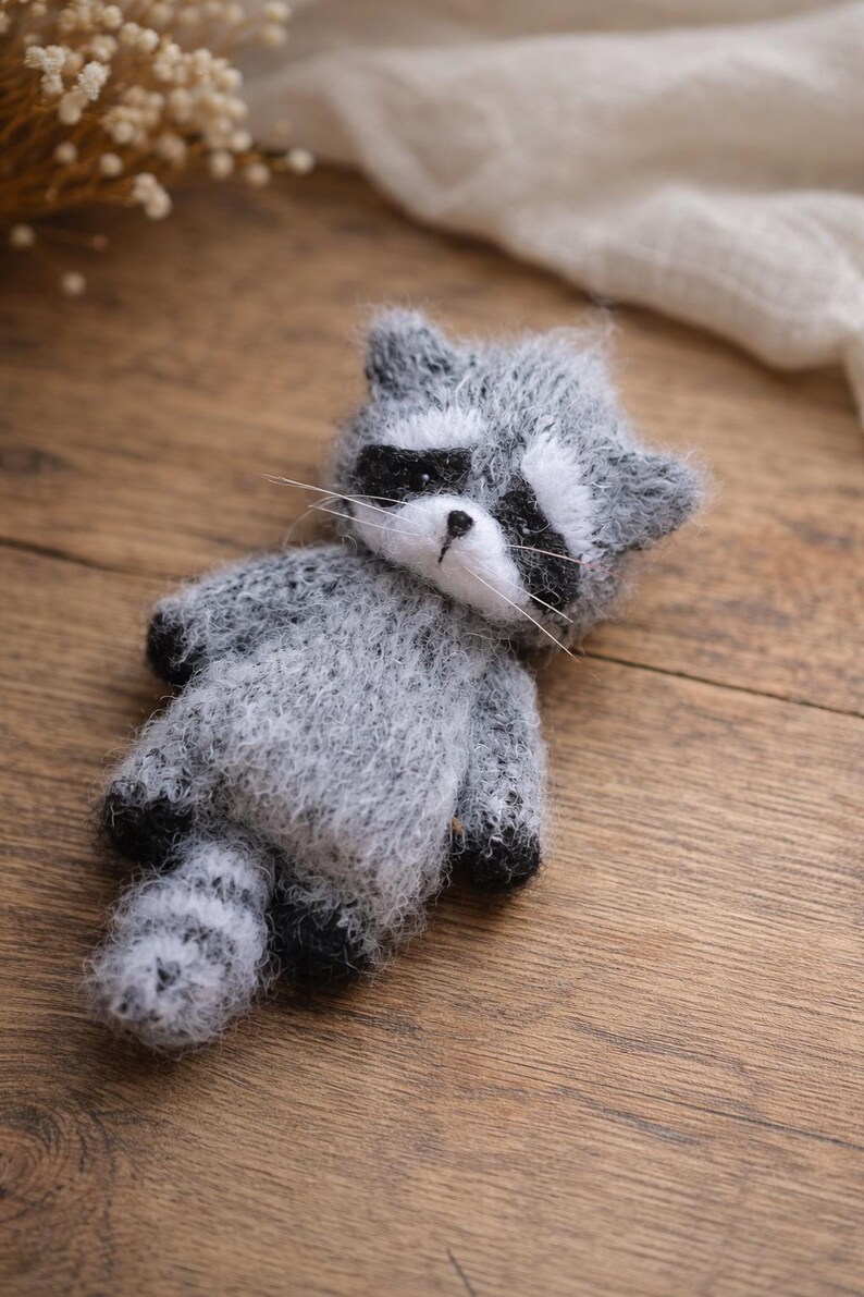 May include: A small, fuzzy, handcrafted raccoon figurine made of grey and white yarn. The raccoon features a distinct black mask around its eyes, a small black nose, white whiskers, and a striped tail. The texture is soft and fibrous throughout.